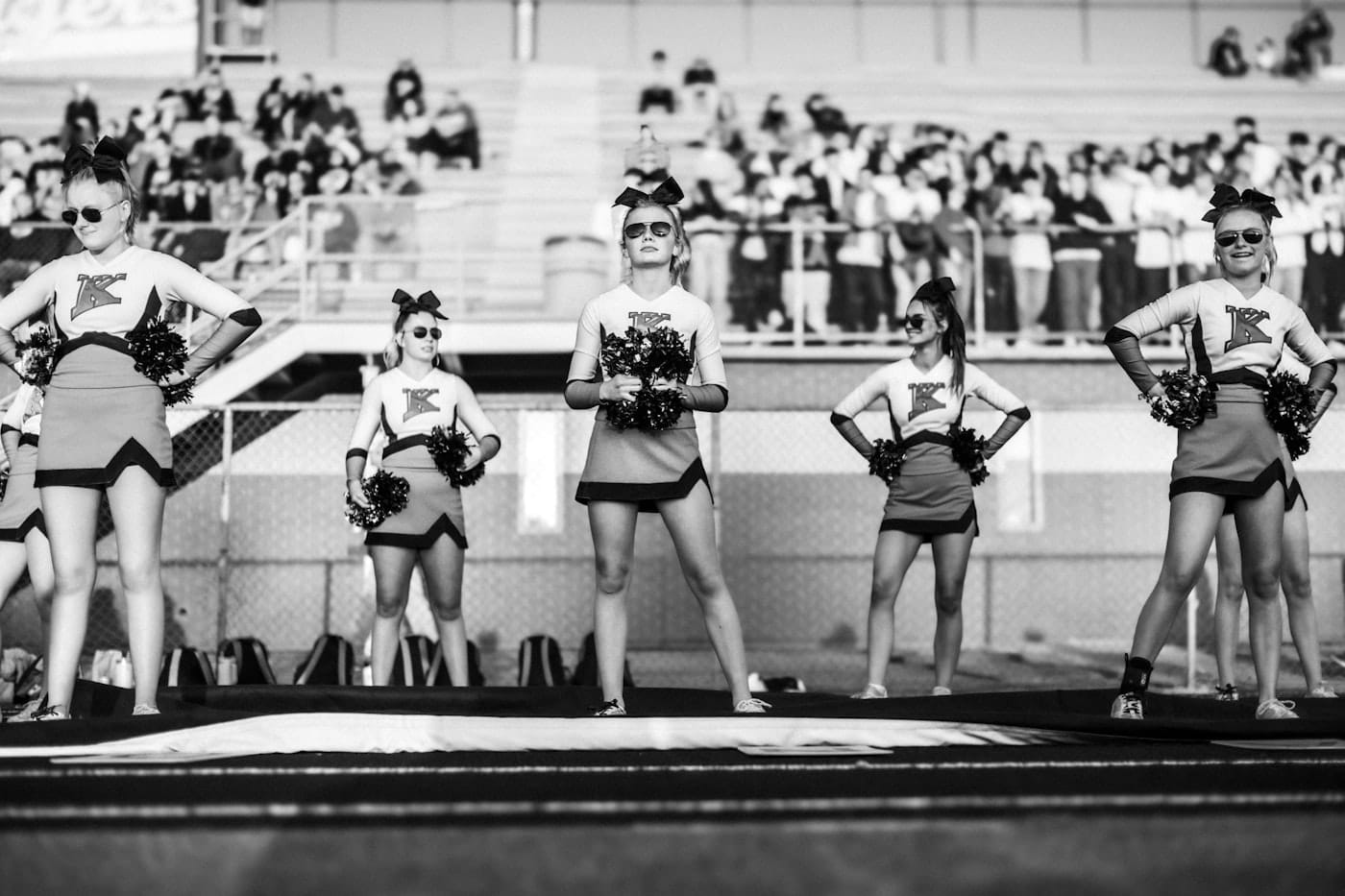 Five cheerleaders pose on a field with spectators behind them